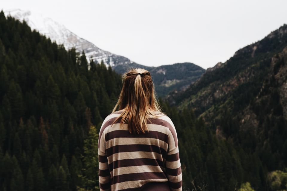 Model in a striped sweater facing pine-covered mountains