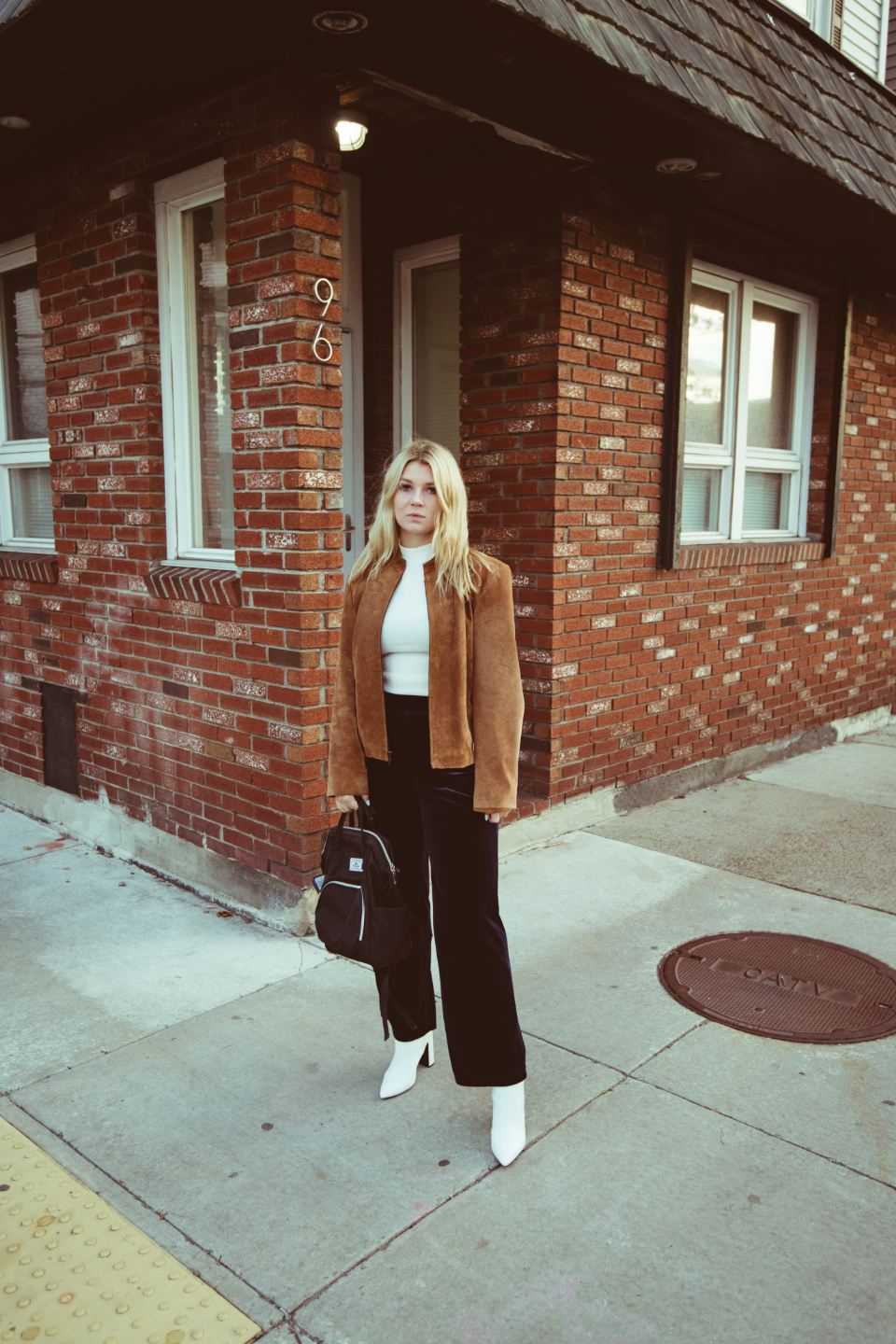 Woman in a camel blazer, white top and black wide trousers by a brick building