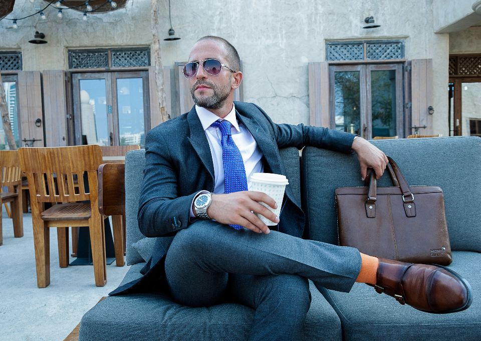 Man in a blue suit and sunglasses seated on a sofa holding a coffee cup