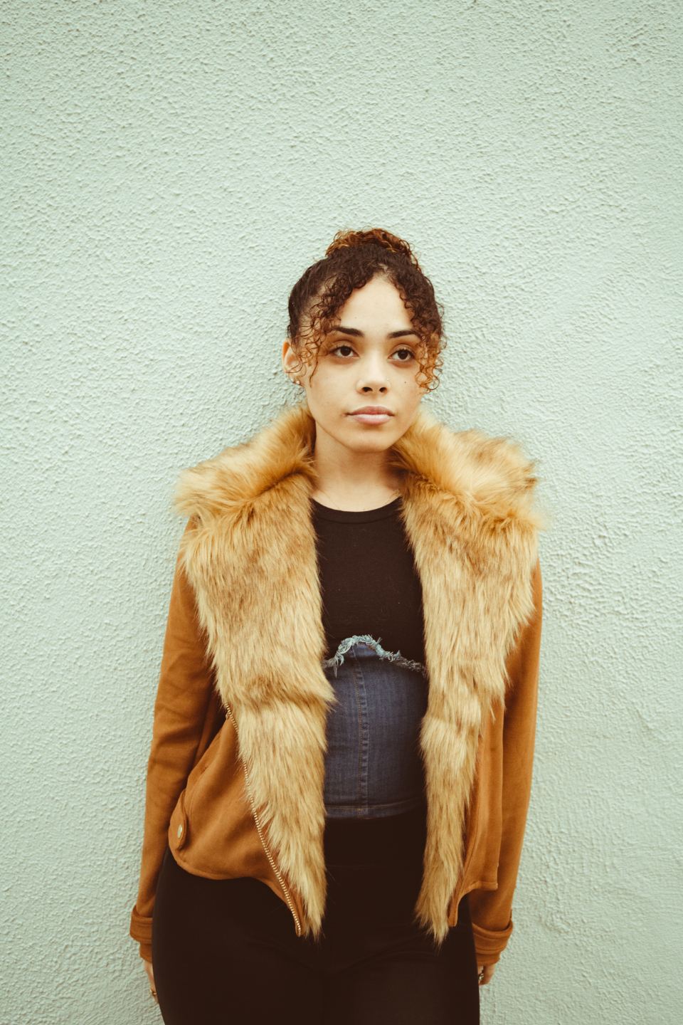 Woman in a tan jacket with a faux-fur collar against a pale wall
