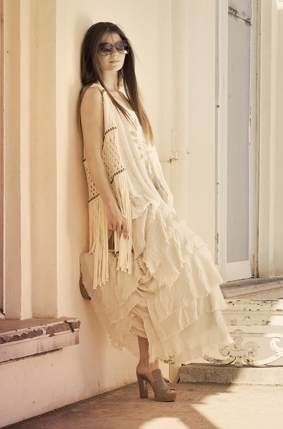 Woman in an ivory tiered ruffle maxi dress and sunglasses leaning against a sunlit cream wall