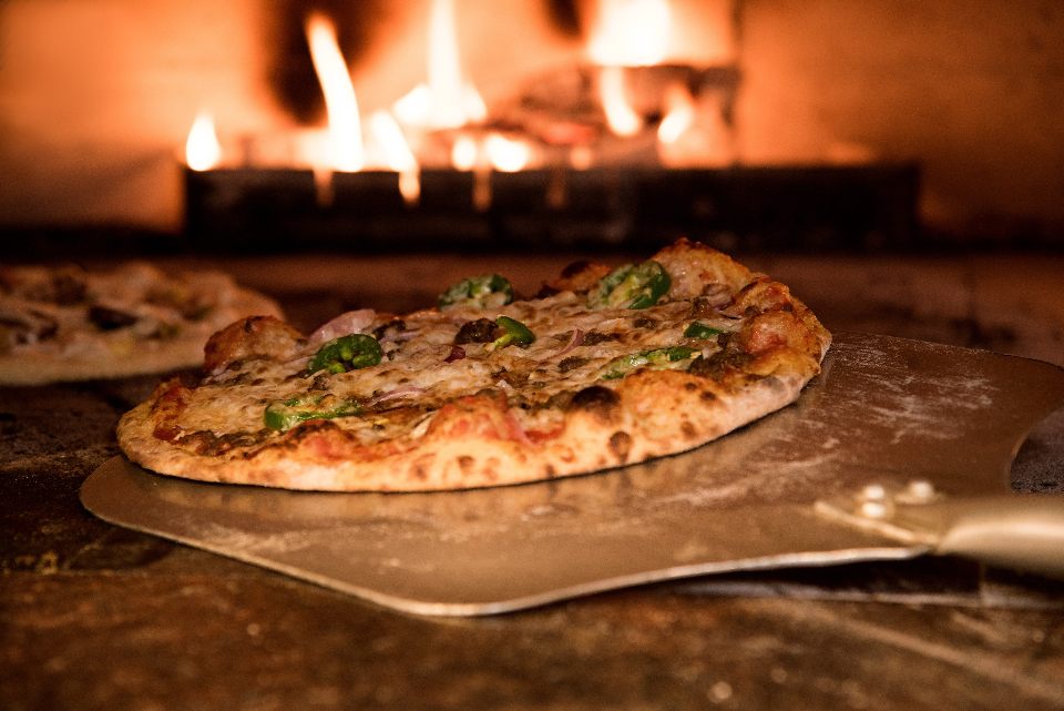 A pizza on a steel peel in front of a burning wood fire