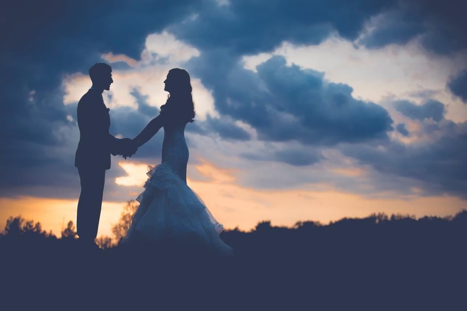 A bride and groom silhouetted against a sunset sky, holding hands